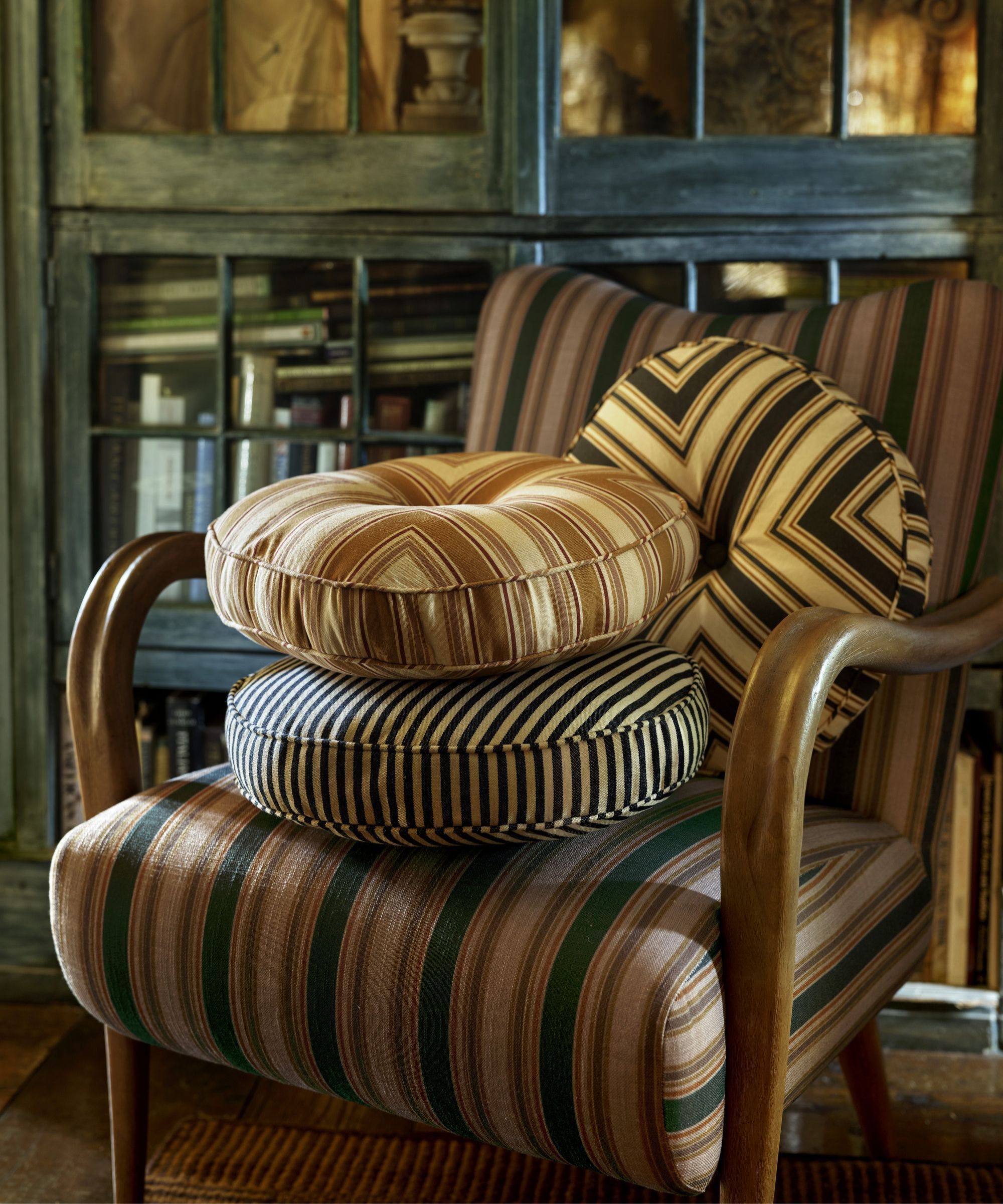 striped round cushions on a striped armchair with brown arms in front of a bookcase filled with books
