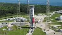 aerial view of a red and white rocket on the launch pad, with greenery and the ocean in the background