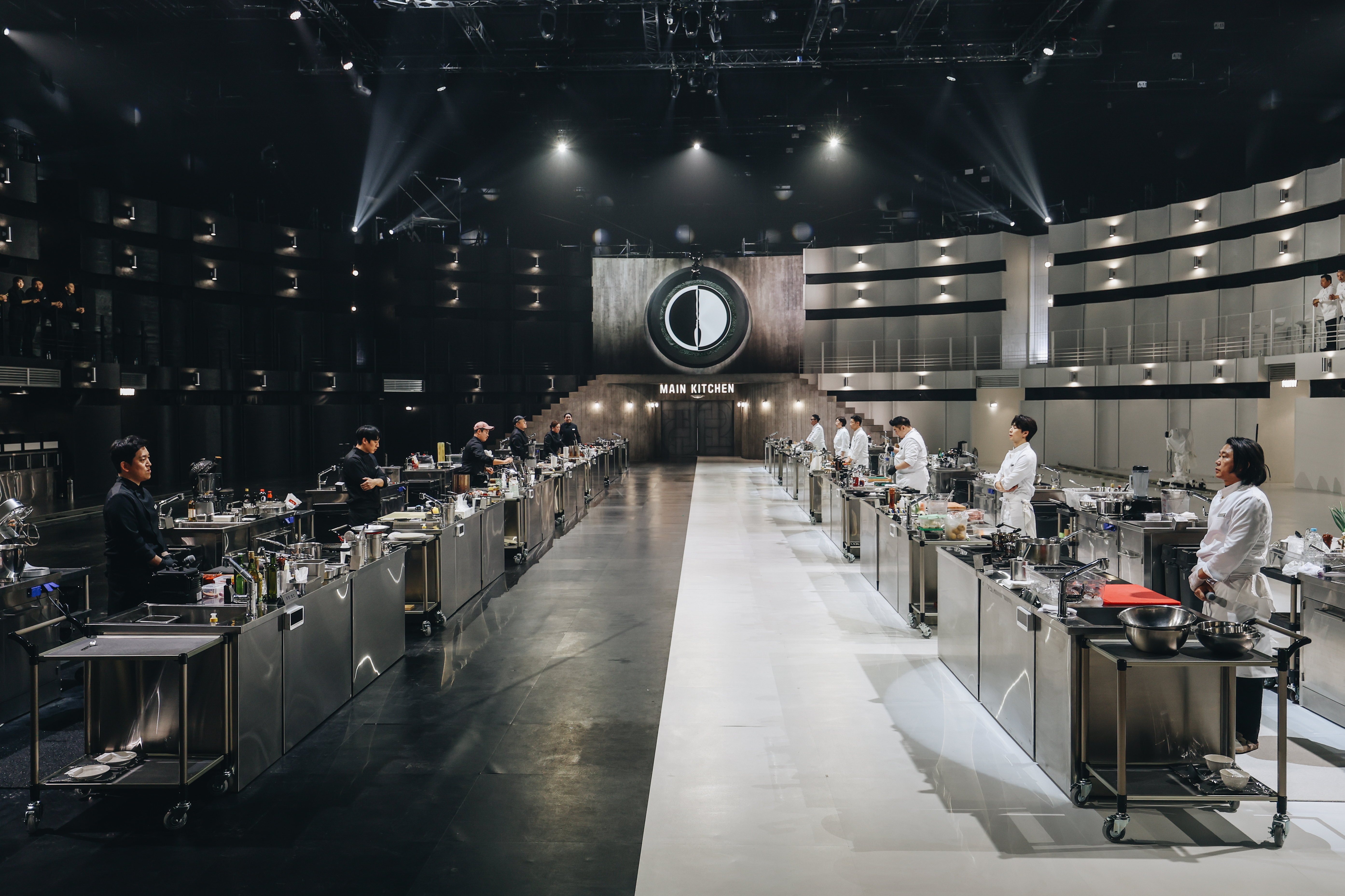 Rows of chefs dressed in black (left) and white (right) stand behind cook stations in a massive set, in a still from 'Culinary Class Wars' season 2.