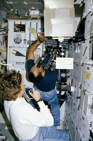 STS-31 mission specialist Kathy Sullivan and pilot Charlie Bolden monitor the student science project "Investigation of Arc and Ion Behavior in Microgravity." In the background can be seen some of the astronauts' personal decals and pennants.