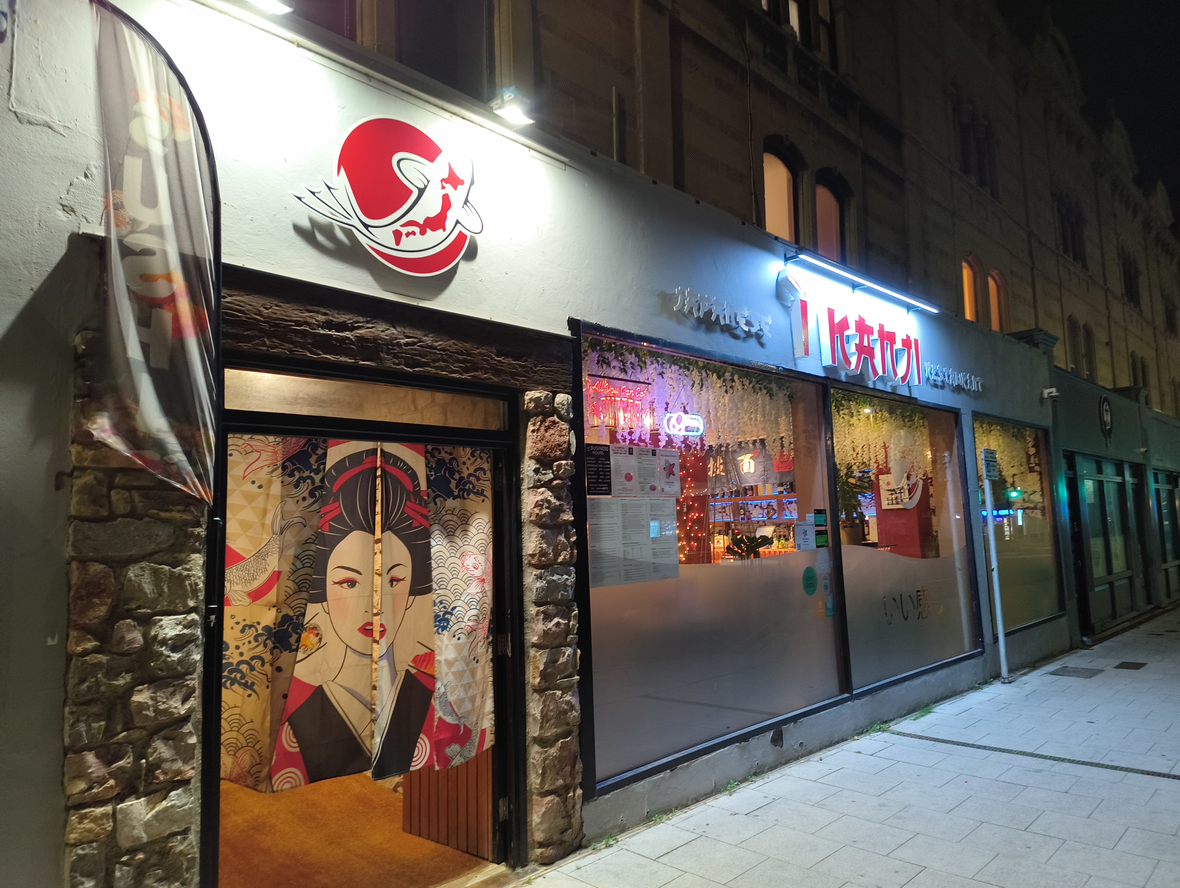 The storefront of a Japanese restaurant called "IKATSU" at night, featuring a curtain at the entrance with a stylized image of a Geisha.