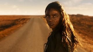 a woman stands in the desert, looking back over her shoulder with a serious expression