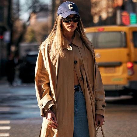 A new york attendee at fashion week fall winter 2025 wearing trench coat and navy baseball cap and sunglasses 