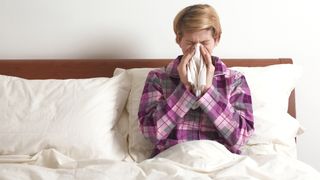 A woman with short blonde hair blows her nose in bed because she has a cold and a stuffy nose
