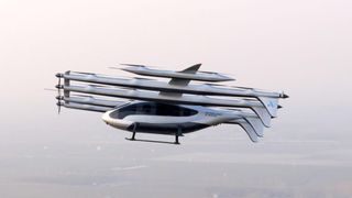 A sleek silvery aircraft flies over a smoggy cityscape with multiple propellors and tails.