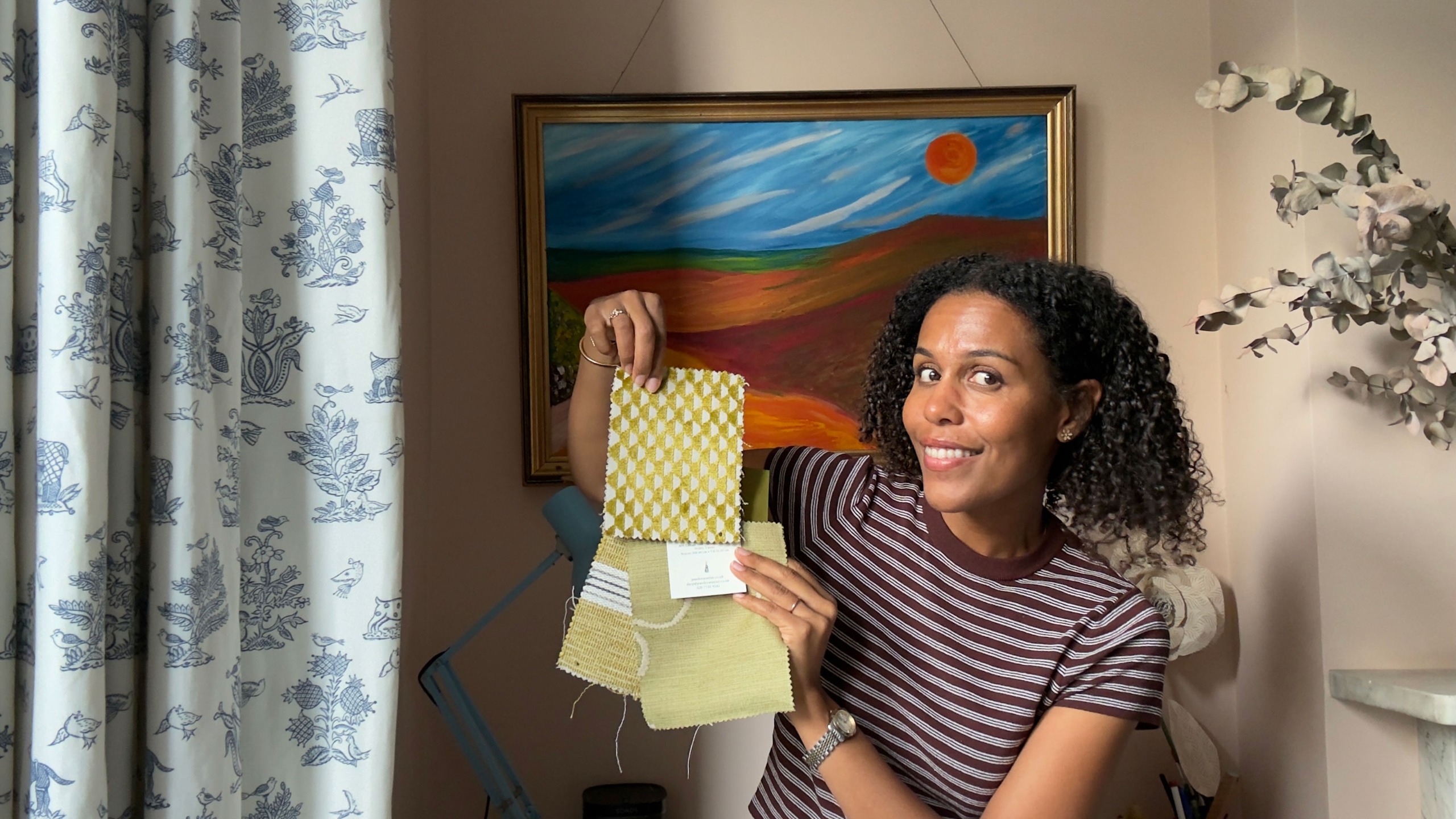 Woman in striped top holding fabric samples, sitting in front of landscape painting