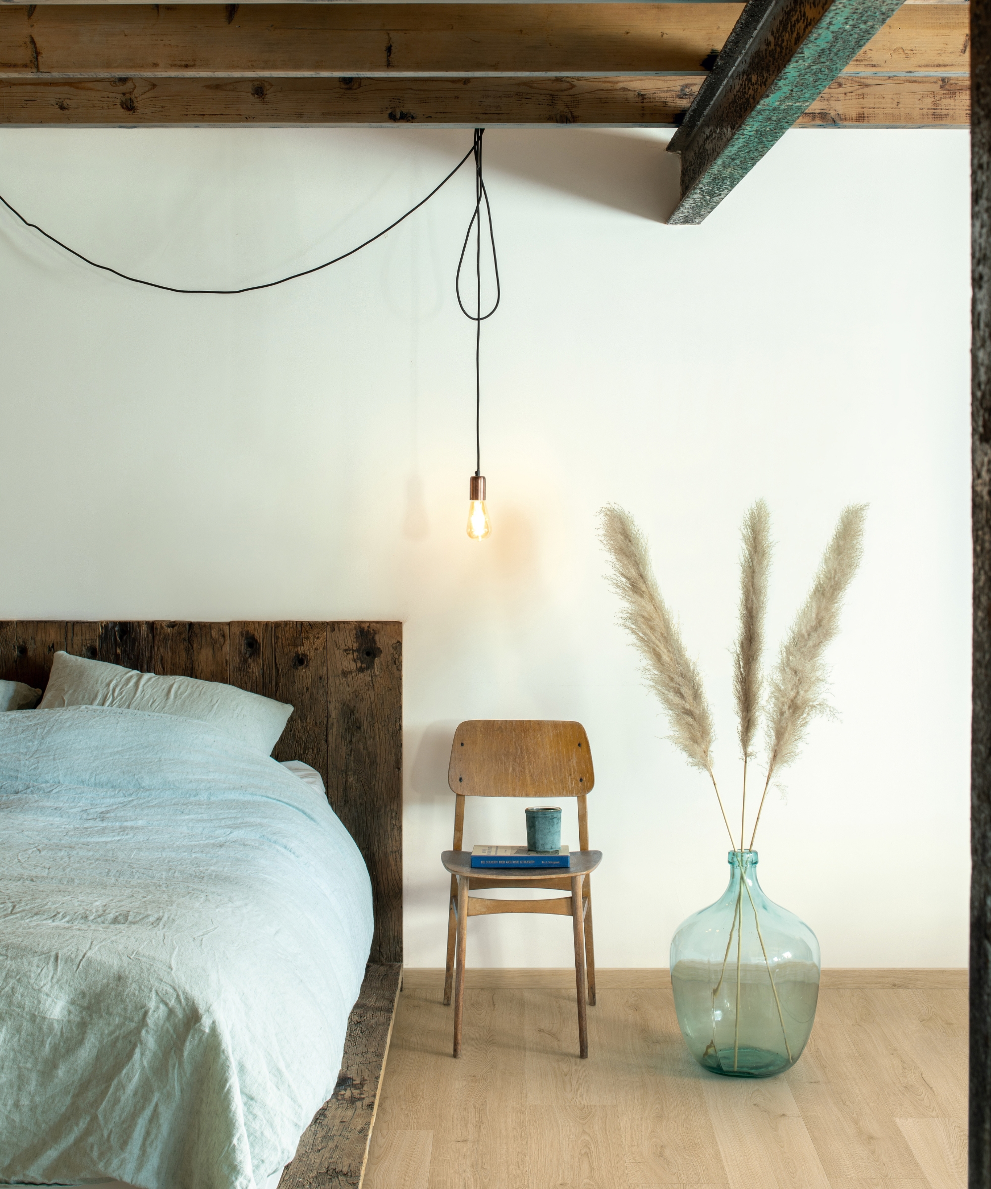 Rustic bedroom with a reclaimed wood bed frame, pale linen bedding, hanging bulb, wooden chair, and pampas grass.
