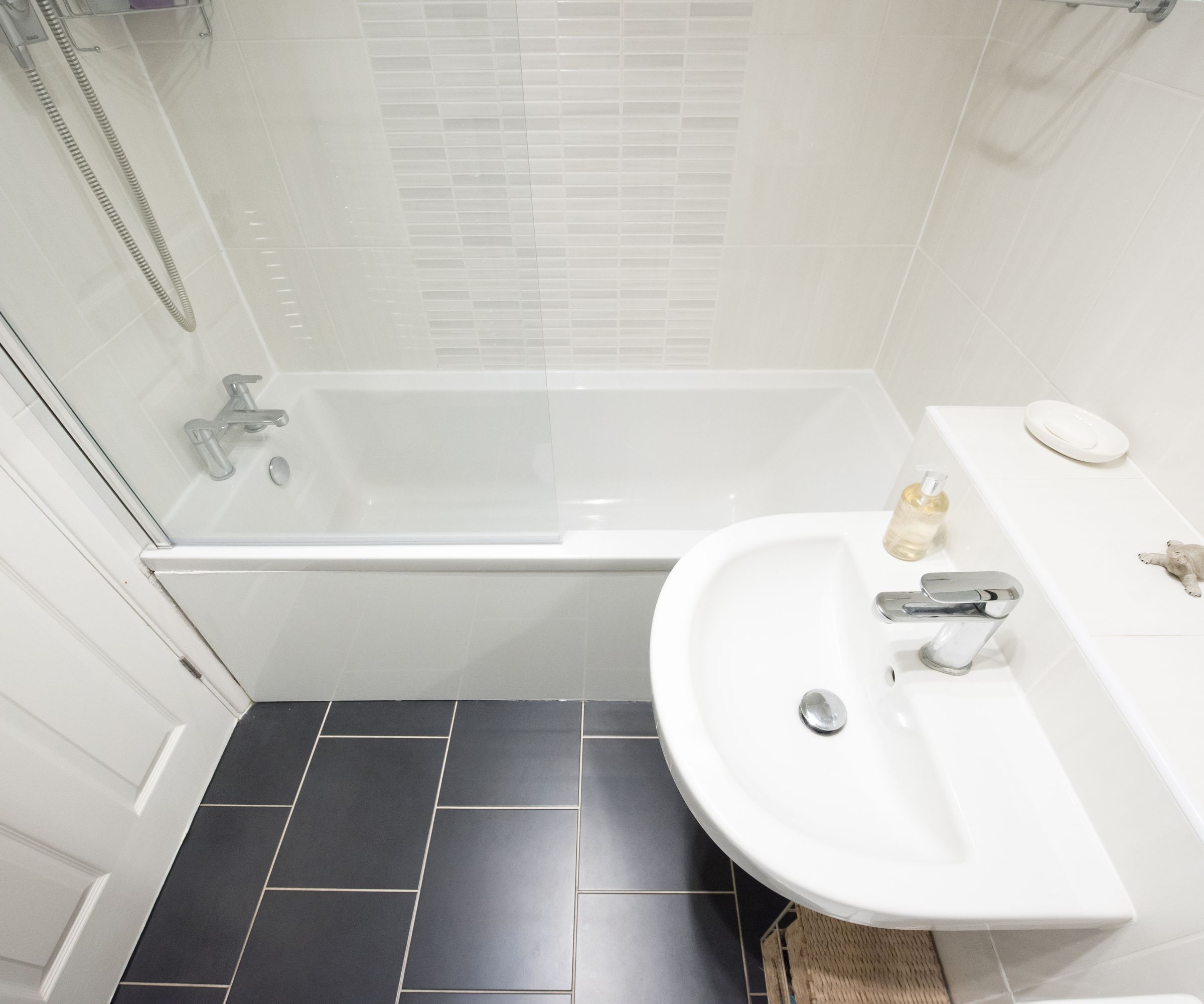 Overhead view of a compact white bathroom with a corner basin, chrome tap, bath with glass shower screen and dark slate floor tiles.