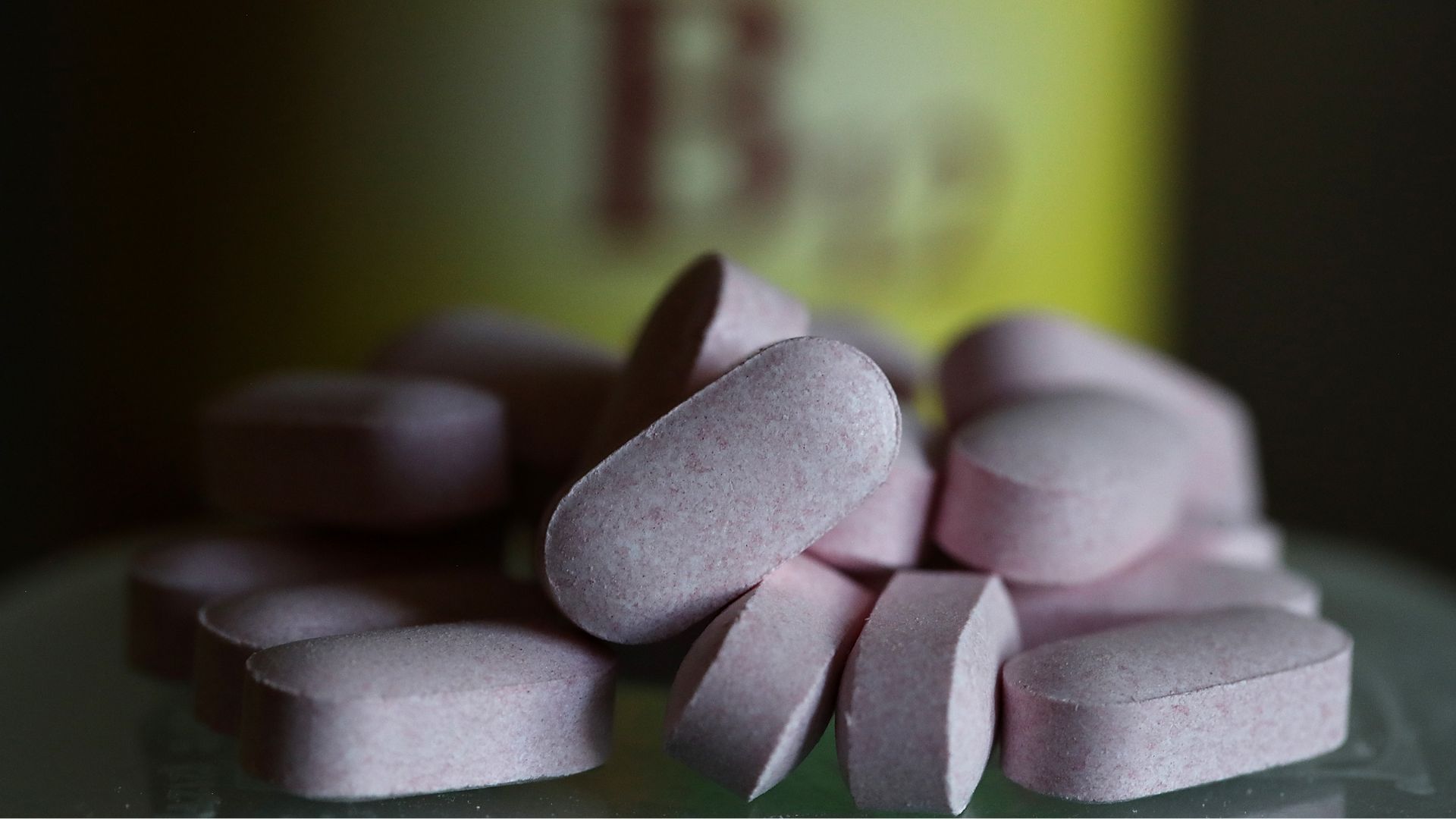 A pile of b12 vitamins in front of an out-of-focus supplement bottle with a yellow label that says &quot;B12&quot;.