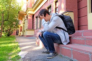 A teenager with a backpack sits outside a home, looking very sad.