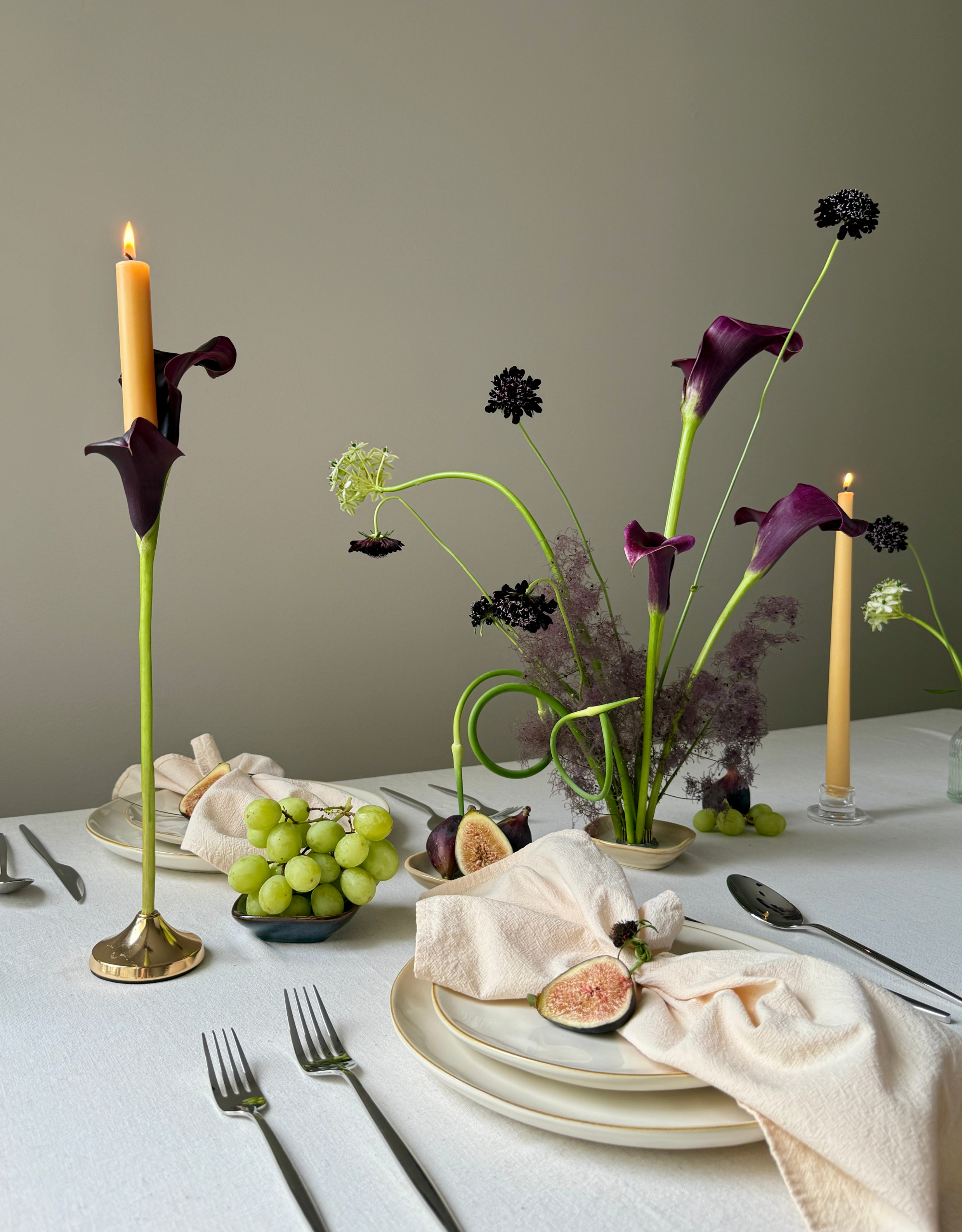 A table set with a calla lily candle stick, a calla lily arrangement, figs, and grapes, beside stacked plates and cutlery