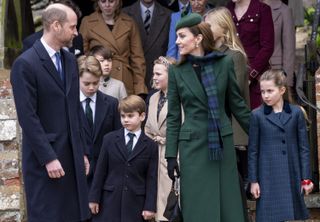 Prince William wearing a blue coat smiling and looking at Kate Middleton, in a green coat and plaid scarf, standing with Prince George, Princess Charlotte and Prince Louis outside church