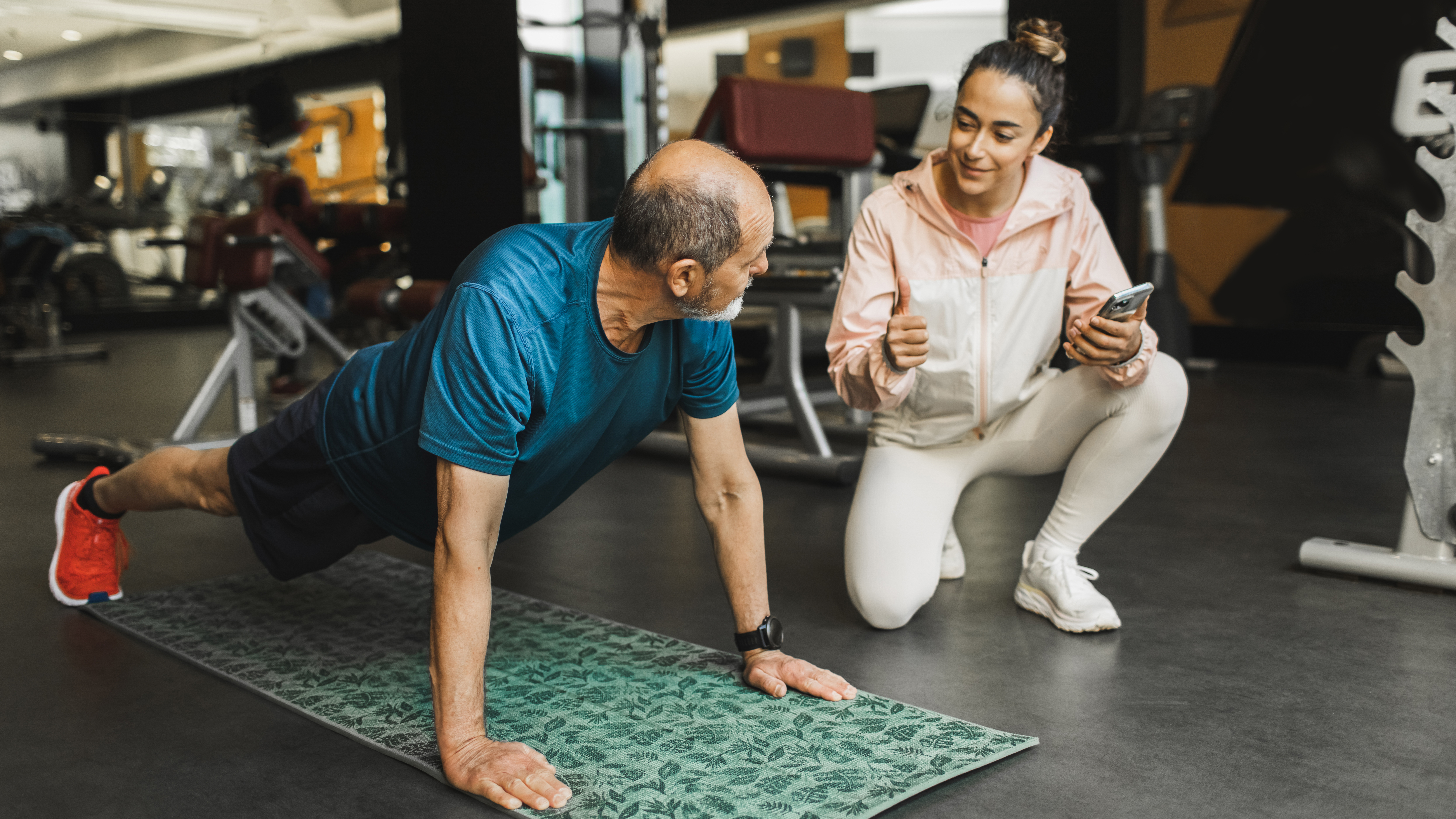 A woman coaches a man through push-ups in a gym. He is looking at her as he exercises on a mat, his hands and toes on the floor, legs and arms straight.