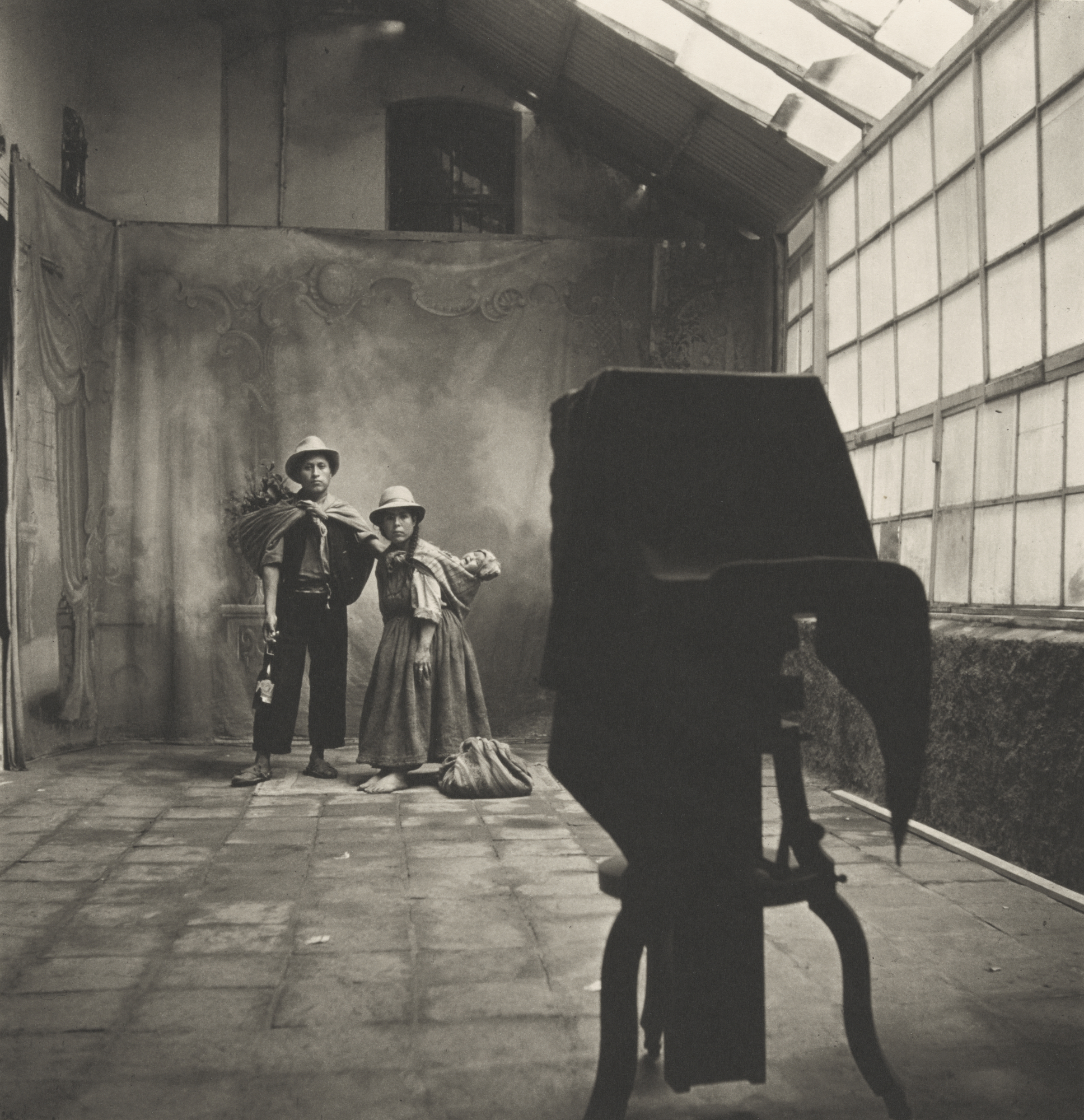 A wide-angle black-and-white photograph by Irving Penn showing a photography studio where two people stand before a painted backdrop, viewed from behind a large, cloth-covered bellows camera.