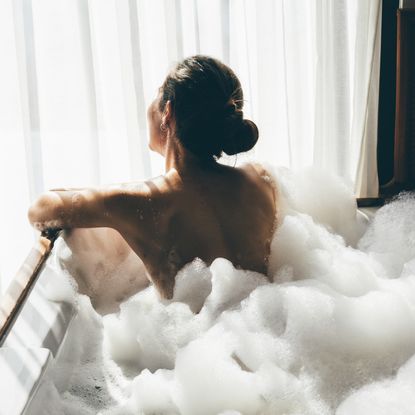 Back view of a woman sitting in a bubble bath facing a window - best bath salts