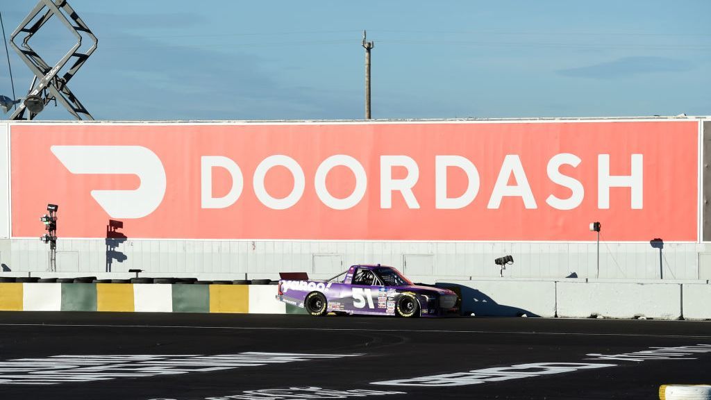 A Nascar racing car driving past a large banner sign displaying the DoorDash logo