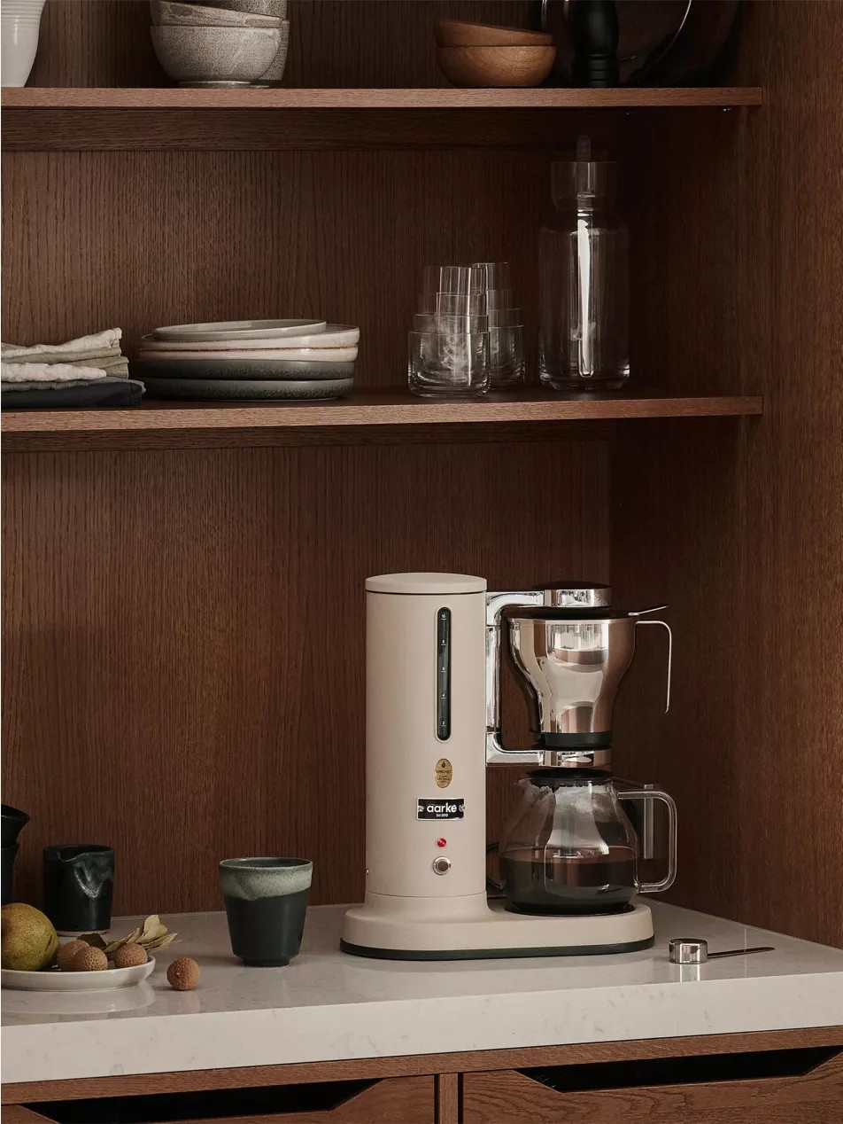 Image of a beige Aarke drip coffee machine on a white counter in a modern, wooden coffee nook.