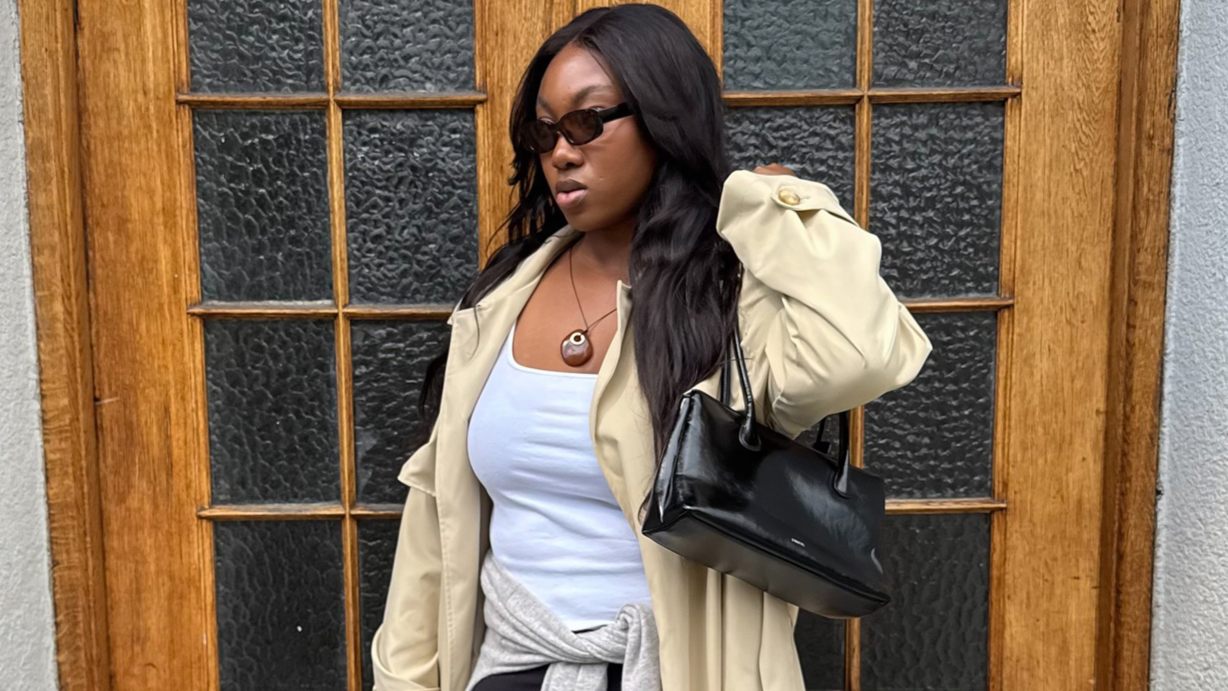 Nnenna wears a trench coat, white tank, black capri pants, a pendant necklace, and a black leather tote.