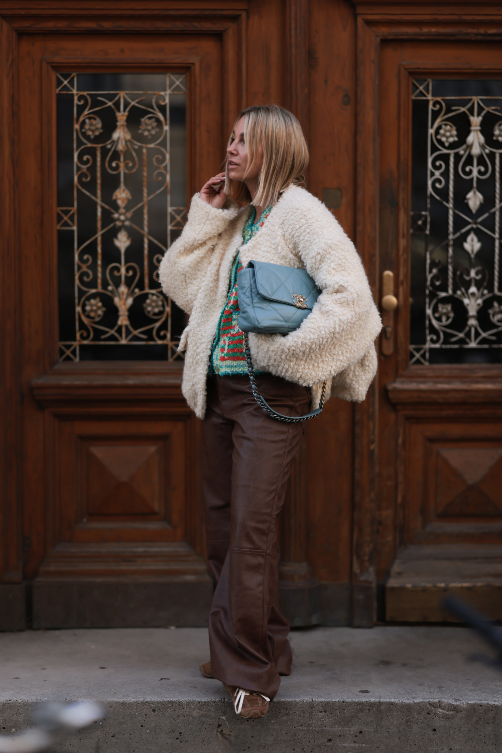 Karin Teigl is seen wearing a yellow gold &amp;ldquo;Alhambra&amp;rdquo; necklace with a green gemstone pendant in a cloverleaf shape from Van Cleef &amp;amp; Arpels; an oversized cream-white fluffy jacket made of mohair and wool with large pockets from Herskind; underneath a multicolored, buttoned wool cardigan with round neckline from Gucci; wide-legged dark brown leather pants from Baum &amp;amp; Pferdgarten; a rectangular blue diamond-quilted leather bag with a gold logo closure and a shoulder strap made of a gold and silver chain interwoven with blue leather from Chanel; and brown suede sneakers with a brown sole and white cotton shoelaces from Miu Miu x New Balance