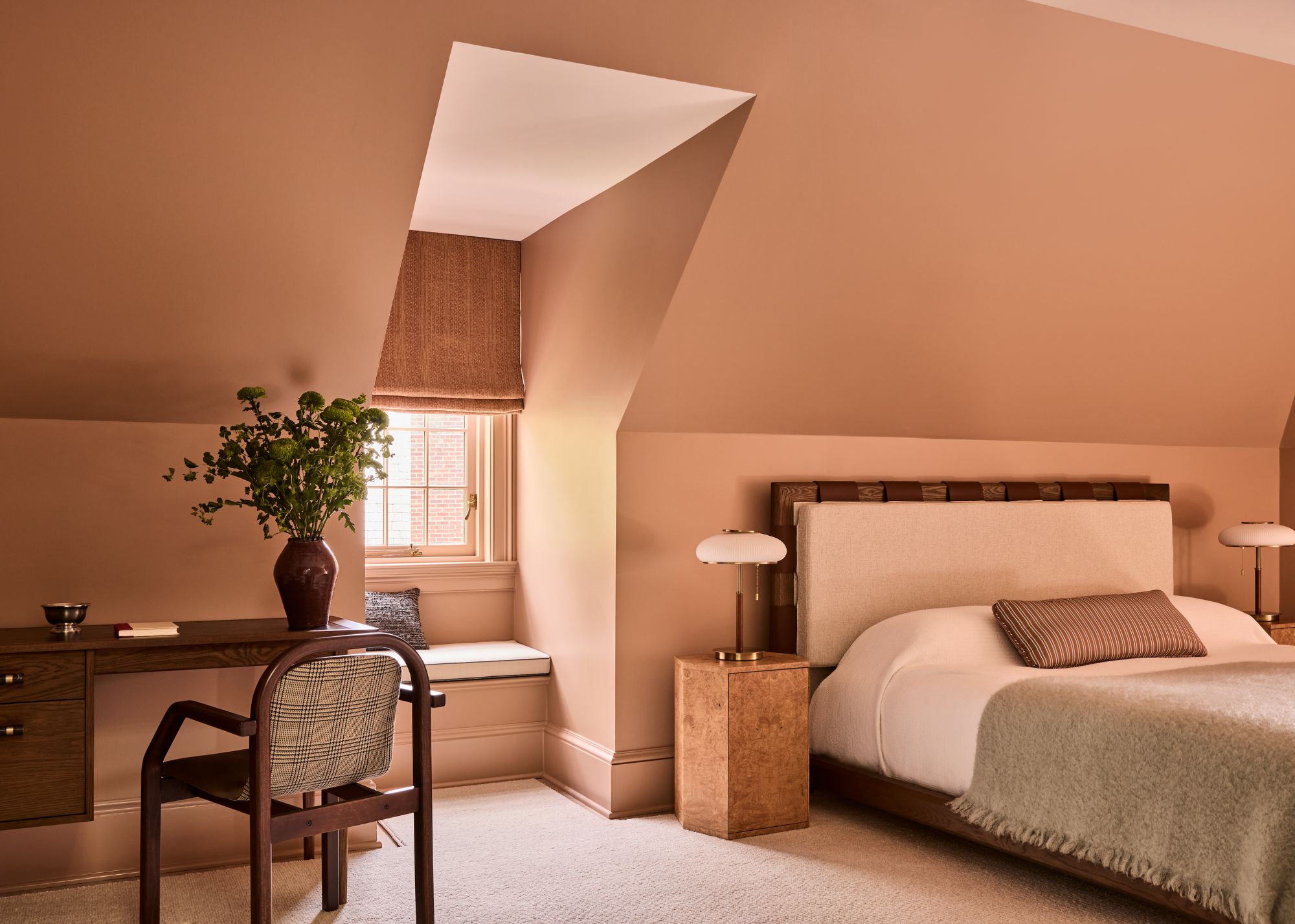 A bedroom with sloped ceilings, color-drenched in a soft shade of pink. A neutral carpet, a low-profile bed with neutral bedding, a wooden desk and chair, and a burl wood nightstand.