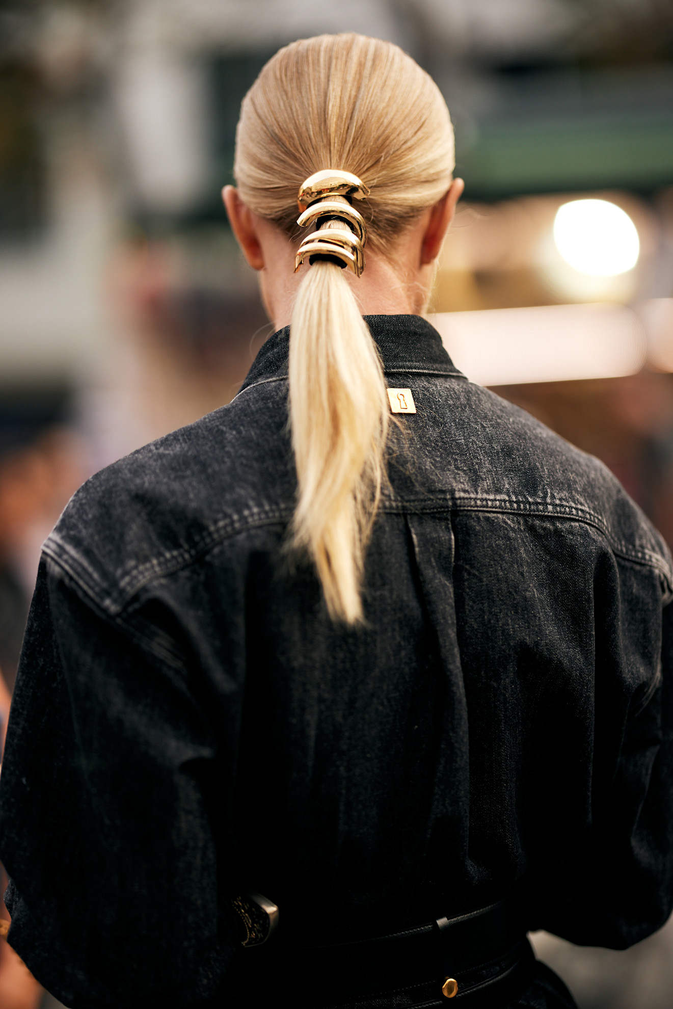 a woman with blonde hair in a ponytail with a gold pony cuff from the back with a black jacket