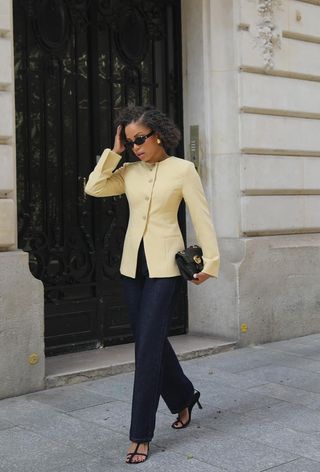 A woman wearing black sunglasses, gold earrings, a yellow blazer, dark-wash jeans, and black t-strap sandals.