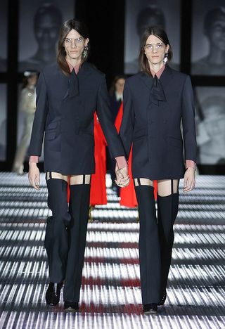 Twin models walking down a runway holding hands wearing dark grey jackets and long dark grey pants which expose their thighs.