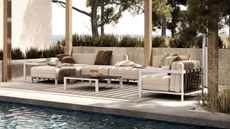 A set of white metal modern outdoor furniture under a wooden trellis on a stone patio, next to a pool.