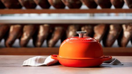 An orange domed bread oven on a wooden counter, with out-of-focus shelves of bread behind it. 