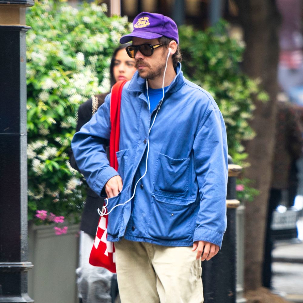 Harry Styled walks down the streets wearing a cobalt blue chore jacket, beige trousers, a purple baseball cap and Maison Margiela Sprinter trainers.