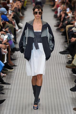 A model at the Miu Miu Spring 2025 show wearing look 15 a navy and grey white breaker and white skirt