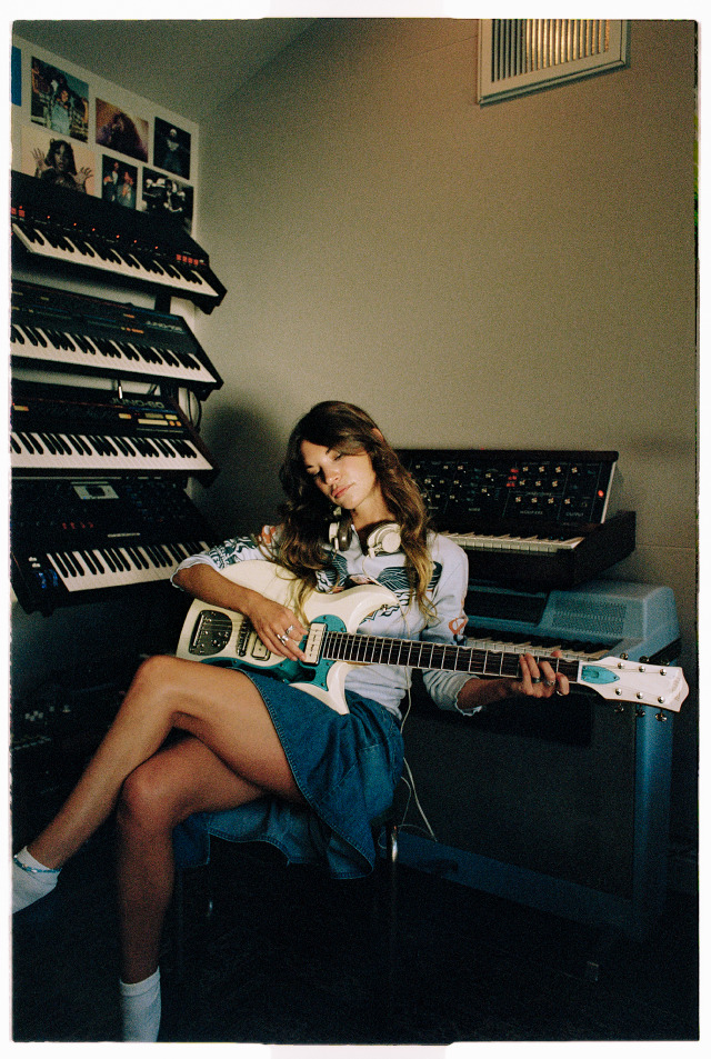 songwriter amy allen sits in a studio playing a guitar surrounded by keyboars and wears a denim skirt and long sleeved shirt