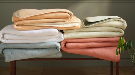 Six folded BrookLinen BreezeWeave collection comforters in assorted colors on a wooden bench, against a moss green wall