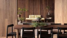 A wooden dining room and living room with a long dining table and black dining chairs. Dried stems in a bowl on the table and potted houseplants around the room