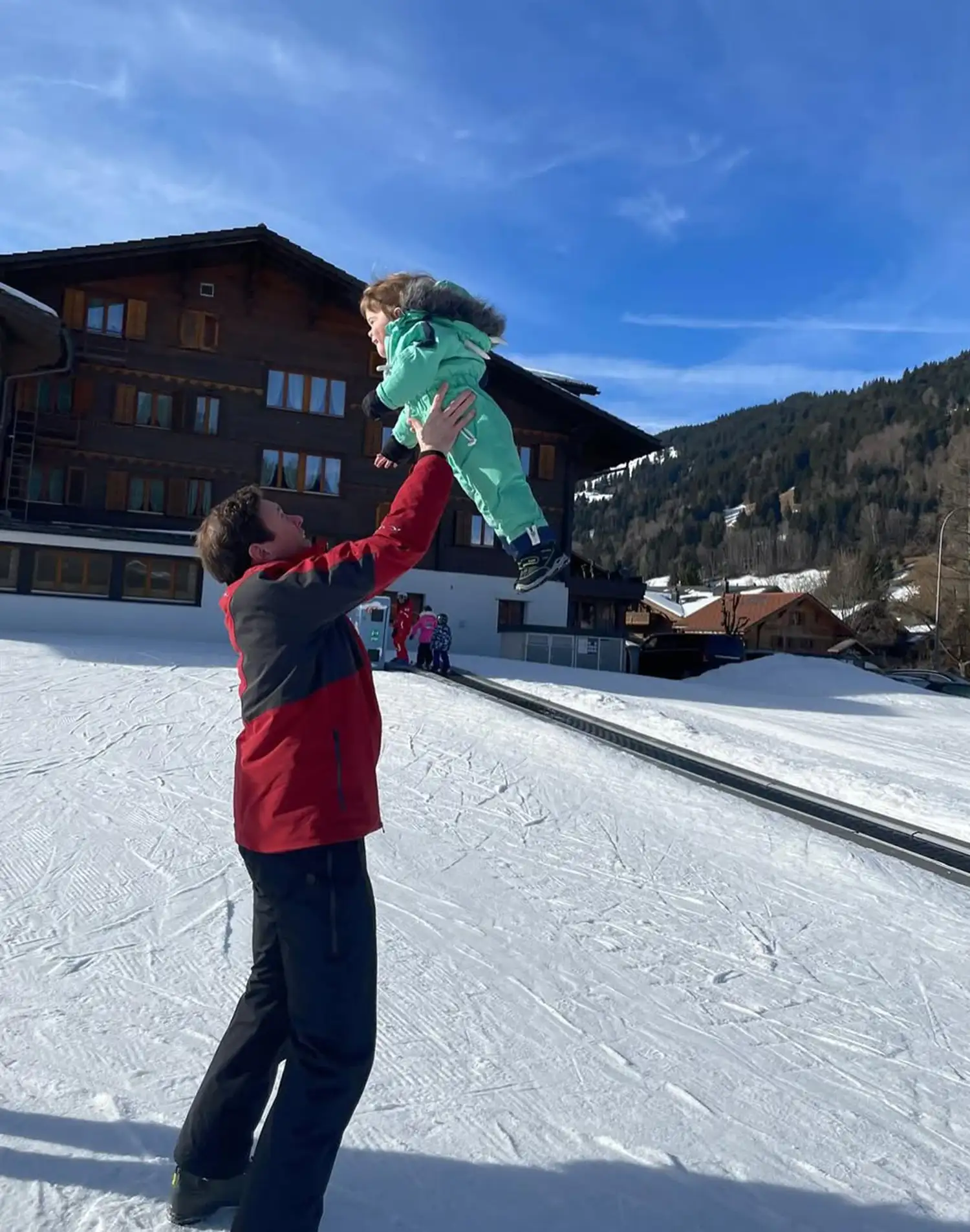 Jack Brooksbank wearing a snow suit and lifting his son in the air in front of a ski resort