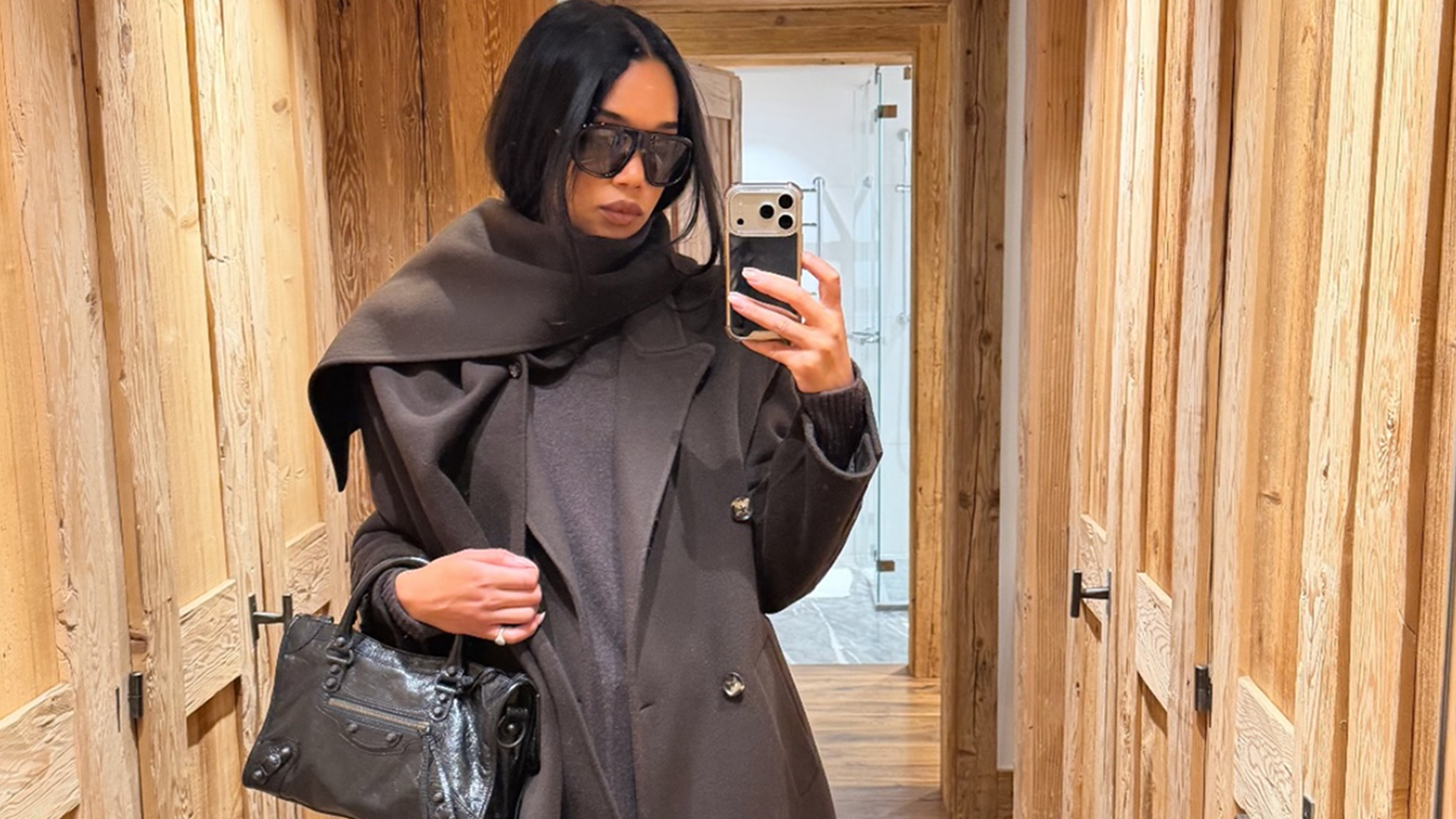 Laura Harrier wearing a brown coat, beige corduroy pants, and brown boots with a black Balenciaga City bag.