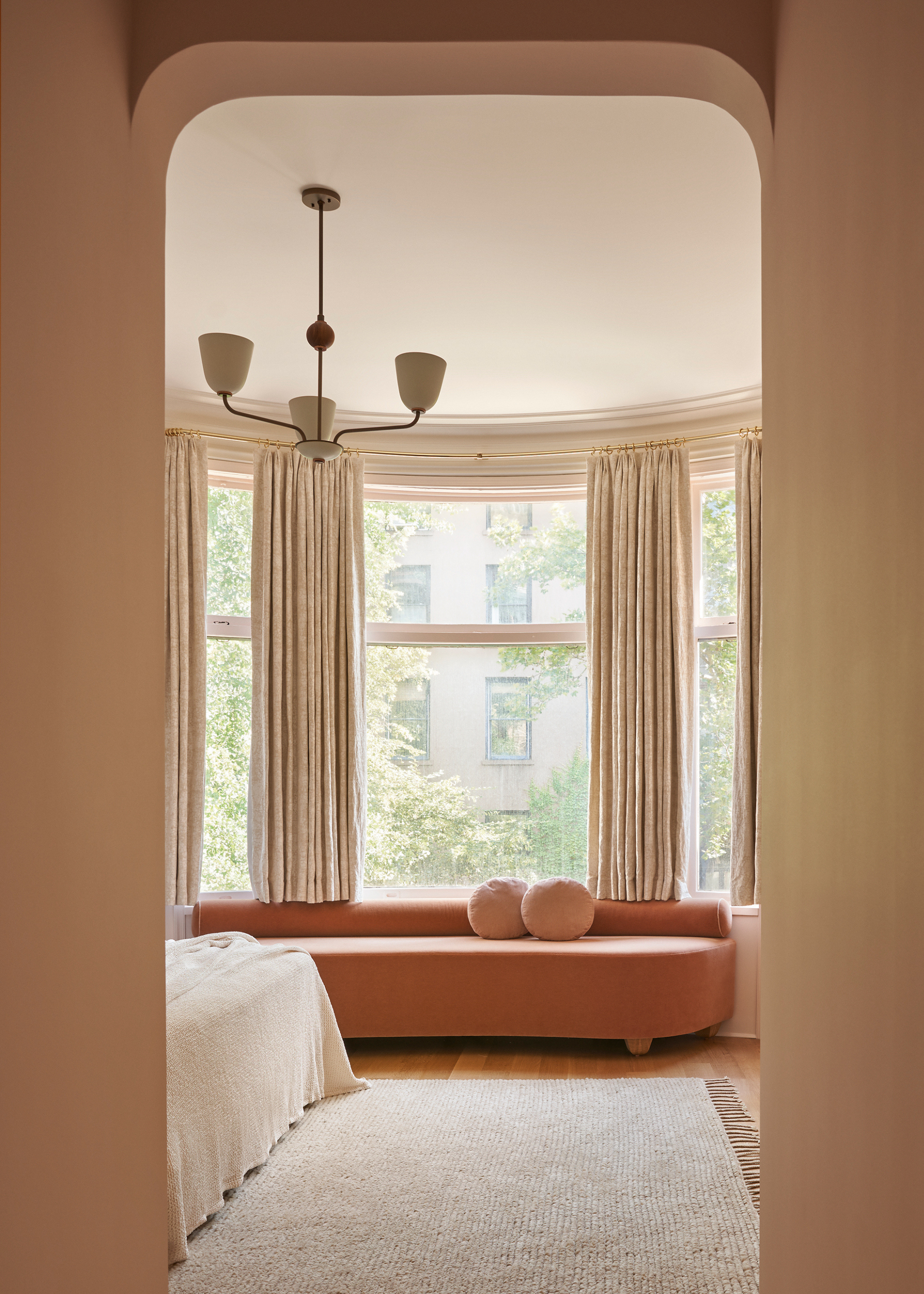 An arched doorway leading to a bedroom with large windows, cream curtains, a soft orange side sofa with a pair of round cushions, a textured rug, and a contemporary pendant light