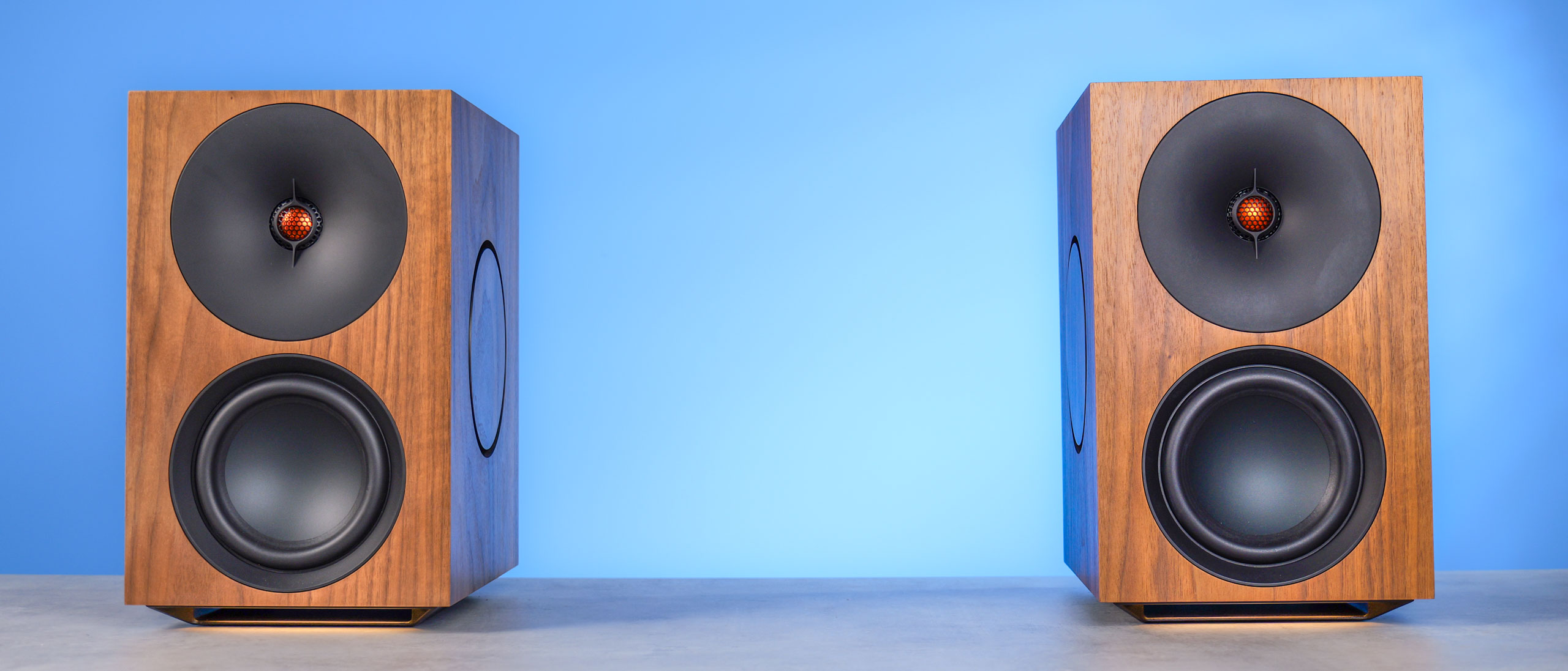 The Cambridge Audio L/R X speakers in walnut color, positioned on a stone surface with a blue background