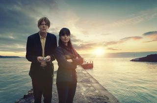 DI Humphrey Goodman (Kris Marshall) and DS Esther Williams (Zahra Ahmadi) stand in front of the ocean in a posed shot, with the sun setting over a breakwater behind them