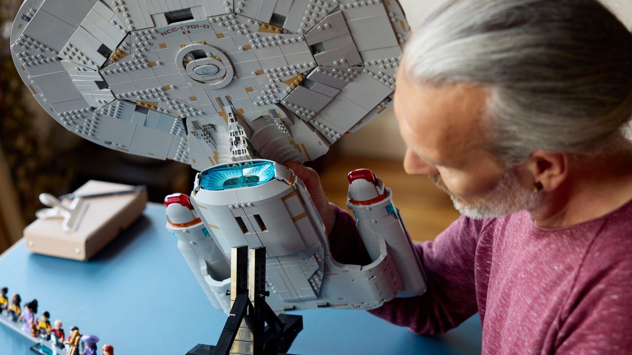A man holds the Lego Enterprise to place it onto a stand, while minifigures can be seen in the background on a blue surface