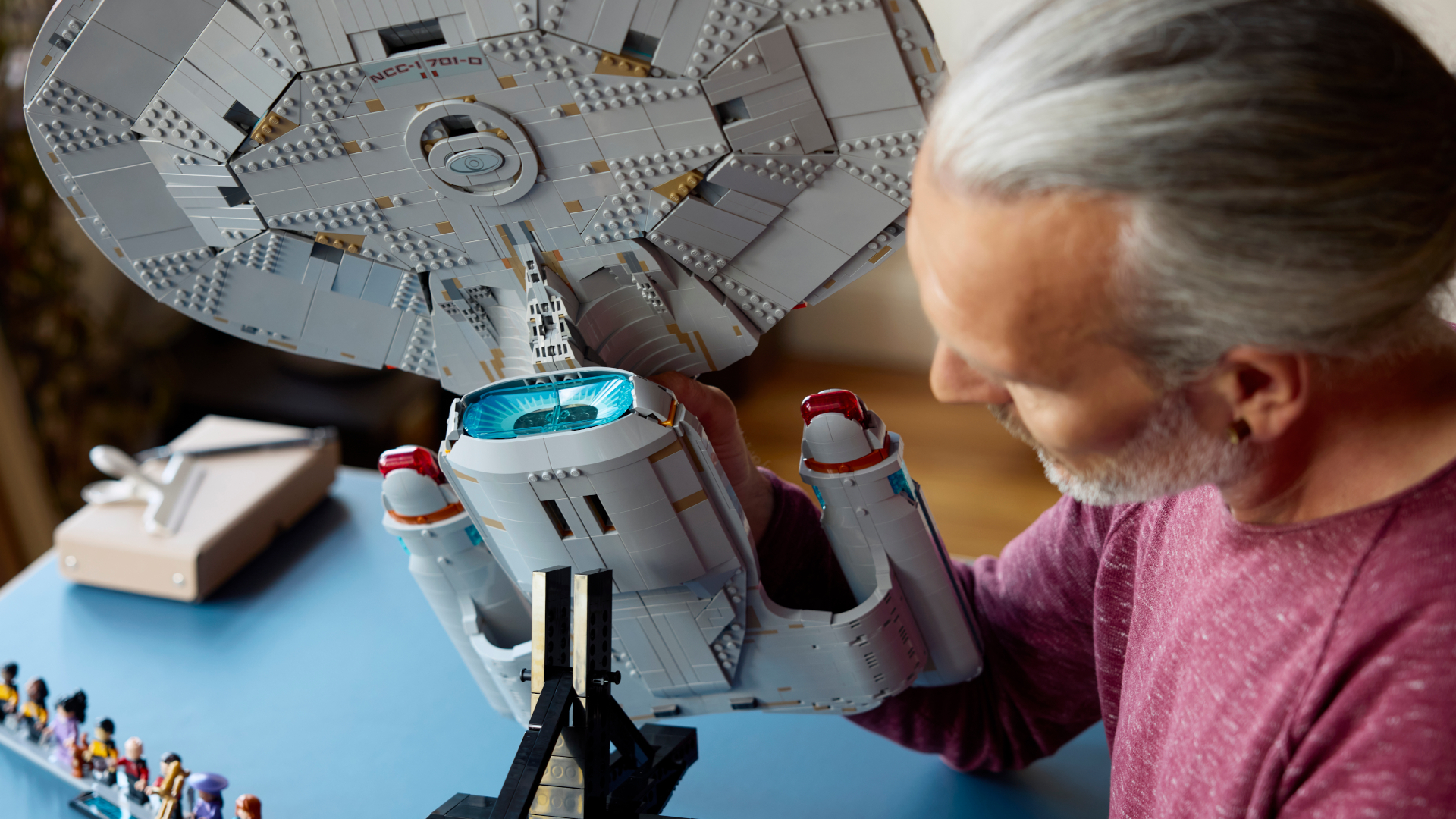 A man holds the Lego Enterprise to place it onto a stand, while minifigures can be seen in the background on a blue surface
