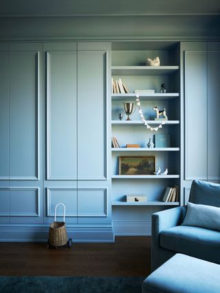 A nursery color drenched in Benjamin Moore's Nimbus Gray blue with built in shelves with decorative accents and cabinetry and an armchair with a pouf and dark wood flooring