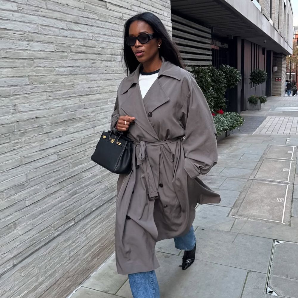 Influencer @nlmarilyn walks down the street in London wearing chisel toe boots with jeans and a grey trench coat.