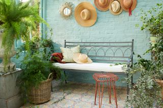 outdoor patio with patterned floor tiles, turquoise painted wall, metal bench, hats on the wall, lush plants