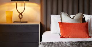 A bedroom by night with beside lamp illuminating a bed with a fabric padded headboard and white sheets layered with sumptuous fabrics and an orange velvet cushion