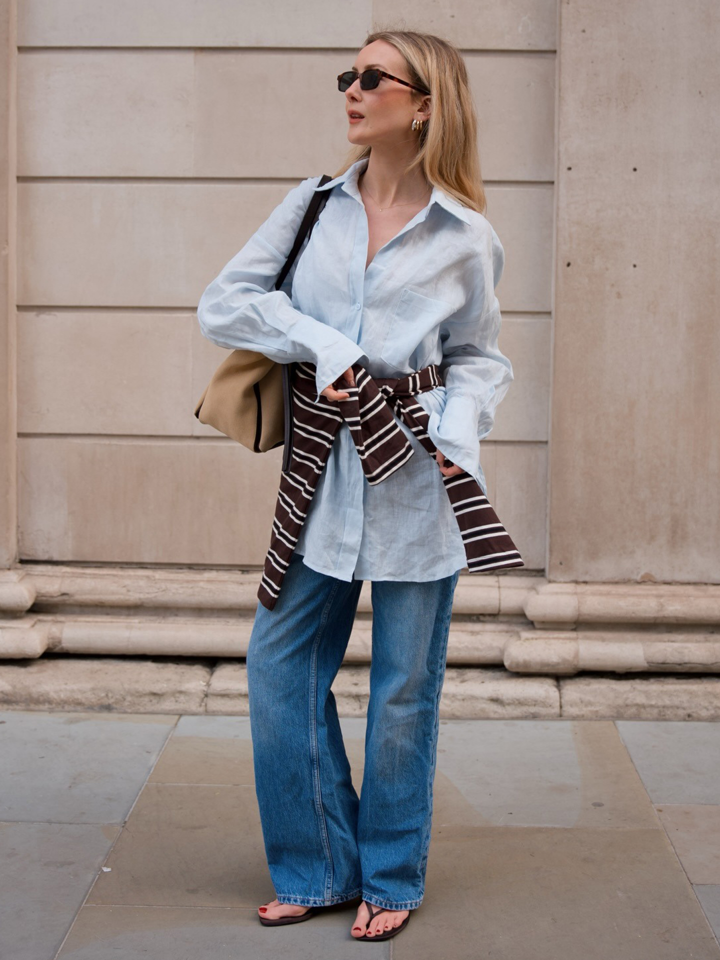 British style influencer Chloe Kath Butler poses in London wearing a long light-blue linen button-down shirt, tan suede bag, striped shirt around the waist, baggy jeans, and brown flip-flip sandals