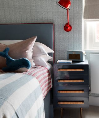 child's bedroom with gray headboard, striped throws, blue toy whale, red wall light and dark gray bedside chest
