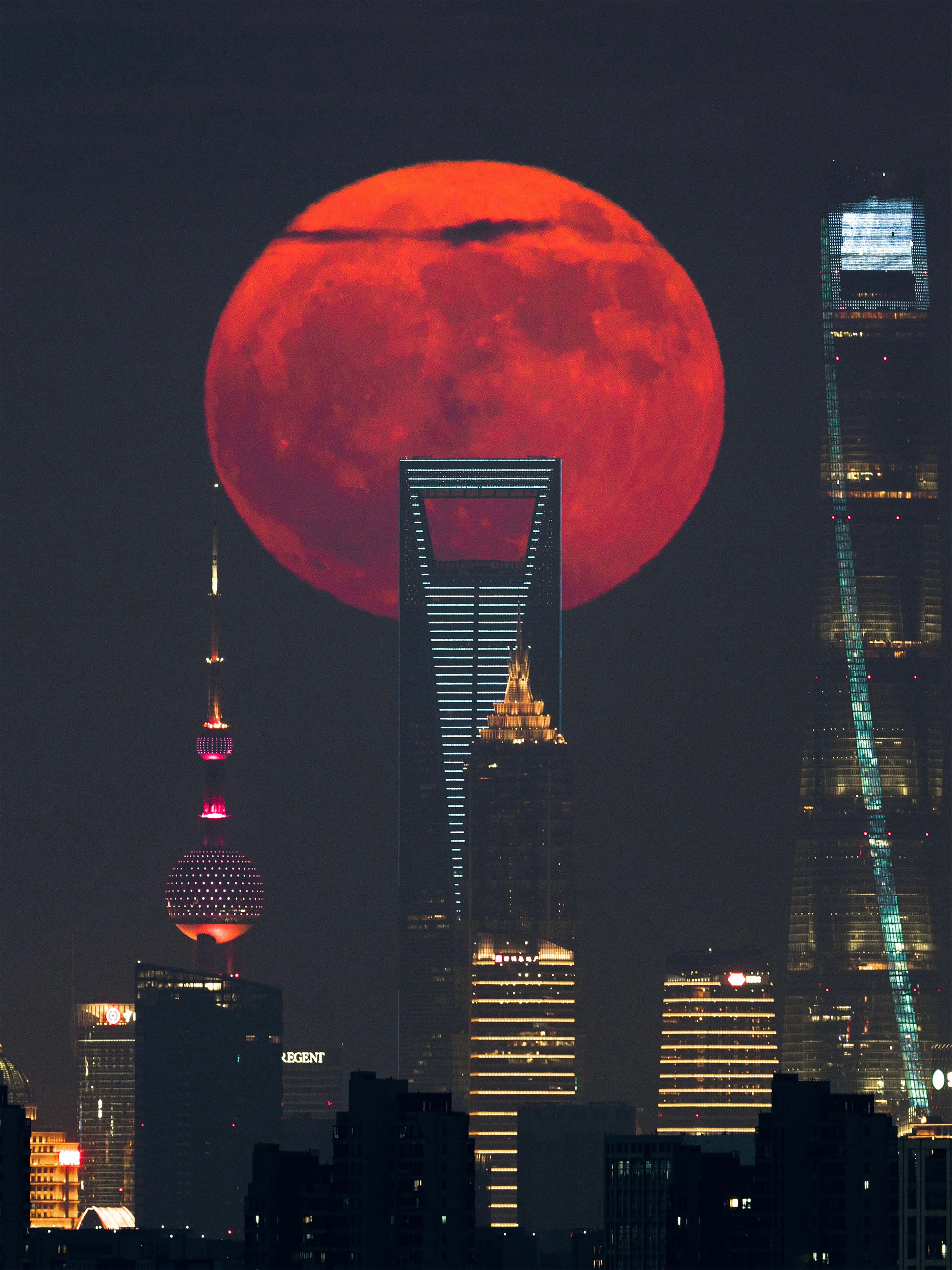 A large bright red moon sits behind a cityscape with tall skyscrapers during the evening