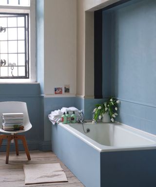 Bathroom with blue tiles, matching walls and tub, vase of white tulips, white chair, small bath mat, wooden floorboards