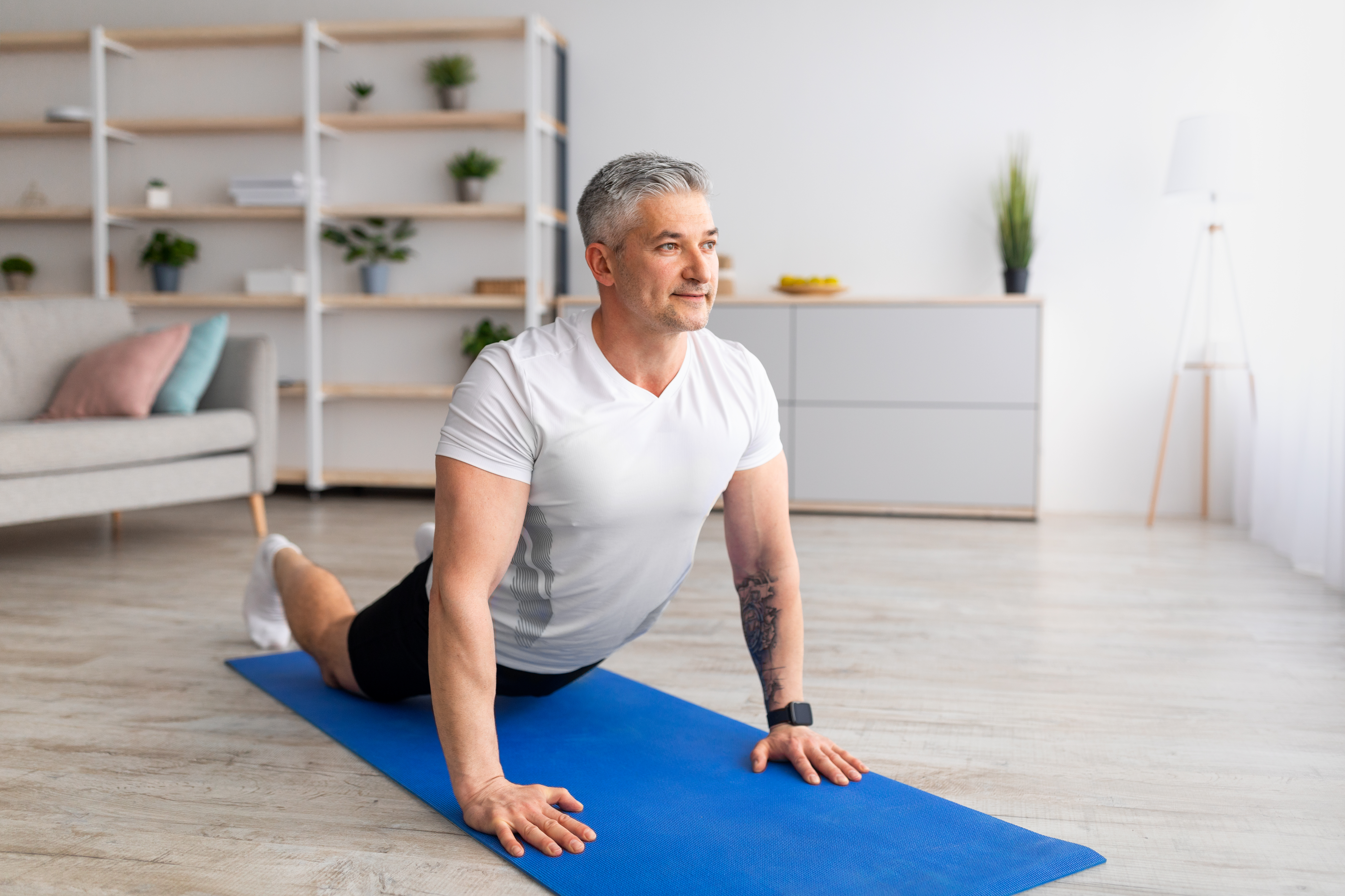 A man practices cobra pose at home on a yoga mat. He is on his front, with his legs long on the mat and his torso held upright by straight arms. Behind him we see a couch and shelves with plants.